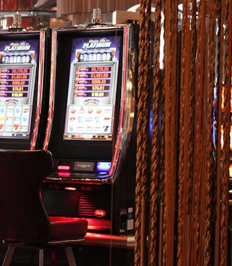 A pair of casino slot machines, each with detailed screens displaying various game outcomes and jackpot amounts. The chairs in front of the slot machines are dark and cushioned for comfort. The background features a hanging curtain made of golden ropes and other casino decorations are visible.
