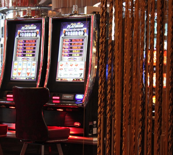 A pair of casino slot machines, each with detailed screens displaying various game outcomes and jackpot amounts. The chairs in front of the slot machines are dark and cushioned for comfort. The background features a hanging curtain made of golden ropes and other casino decorations are visible.