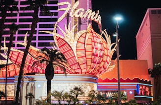 A brightly lit casino with neon lights in vibrant red, pink, and blue, featuring a large flamingo signage. Palm trees are visible in the foreground with vehicles passing by on the street at night.