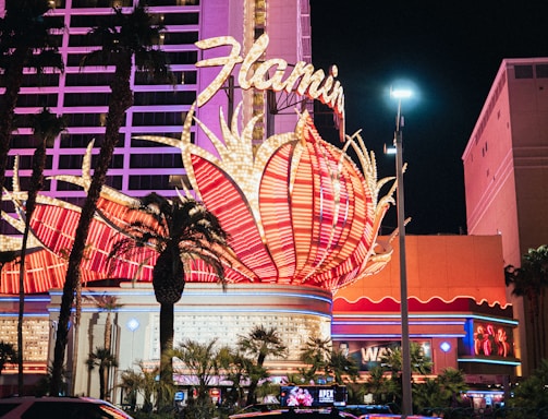 A brightly lit casino with neon lights in vibrant red, pink, and blue, featuring a large flamingo signage. Palm trees are visible in the foreground with vehicles passing by on the street at night.